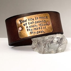 May include: A brown leather bracelet with a brass plate engraved with the quote "Your life is made of two dates & a dash. Make the most of the dash."