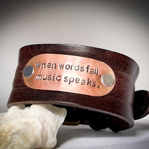 May include: A brown leather bracelet with a copper plate that reads "when words fail, music speaks."