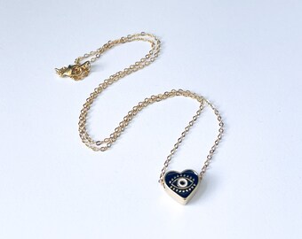 May include: A gold chain necklace with a heart-shaped pendant featuring a black enamel evil eye design.