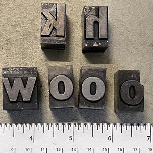 May include: A set of metal letterpress printing blocks spelling out the word "WOOD". The blocks are made of metal and have a worn, vintage look. The letters are raised and ready to be inked for printing.