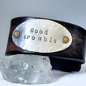 May include: A brown leather cuff bracelet with a silver oval plate that says "good trouble". The plate is hammered and has two brass rivets.