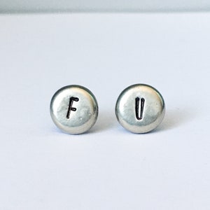 May include: A pair of silver-colored stud earrings. One earring has the letter "F" stamped on it, and the other has the letter "U". The earrings are round and have a slightly domed surface.