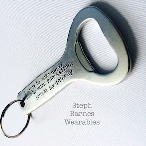 May include: Silver bottle opener keychain with the quote "I drink to make other people more interesting. Ernest Hemingway" engraved on it. The keychain is branded with "Steph Barnes Wearables".