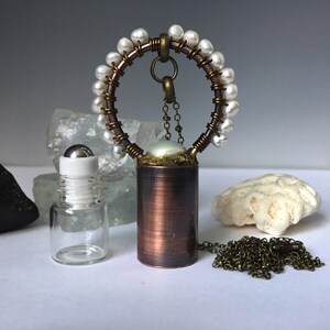 May include: A copper and brass necklace with a pearl pendant and a circle of white pearls. The necklace is on a white surface with a clear glass bottle and a white coral.