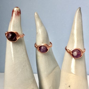 May include: Three copper wire wrapped rings with amethyst gemstones. The rings are displayed on white ceramic ring holders.