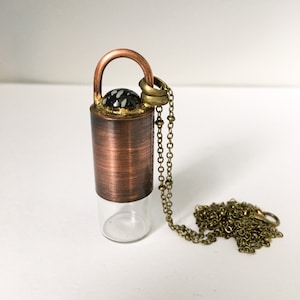 May include: A copper and brass necklace with a clear glass vial pendant. The vial has a copper top with a black and white stone inlay. The necklace is attached to the vial with a brass chain.