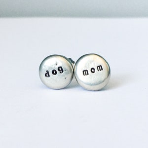 May include: A pair of silver stud earrings. One earring has the word "dog" stamped on it, and the other has the word "mom". The earrings are round and have a hammered texture.