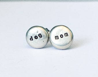 May include: A pair of silver stud earrings. One earring has the word "dog" stamped on it, and the other has the word "mom". The earrings are round and have a hammered texture.
