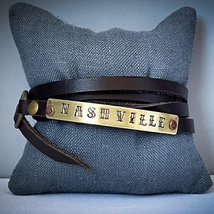 May include: A brown leather wrap bracelet with a brass plate engraved with the word "NASHVILLE".