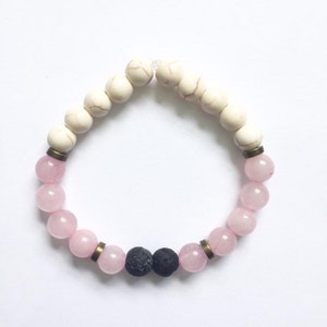 May include: A white and pink beaded bracelet with two black lava beads. The bracelet has gold-toned metal accents.