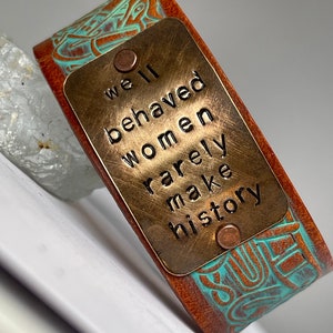 May include: A brown leather bracelet with a turquoise design and a brass plate that reads "Well behaved women rarely make history."