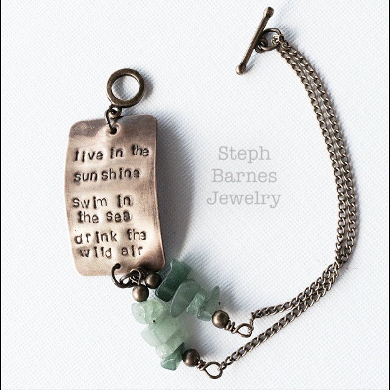 Op de afbeelding: Een koperen armband met een bedel waarop staat "live in the sunshine, swim in the sea, drink the wild air". De bedel is bevestigd aan een ketting met een schuifgesp. De armband is versierd met groene edelsteen kralen.