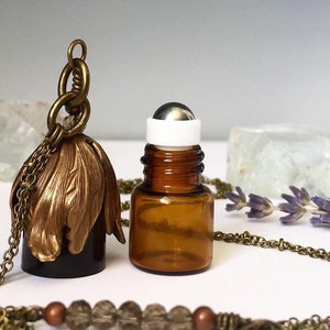 May include: A gold chain necklace with a brown glass bottle pendant and a separate brown glass roller bottle with a silver roller ball. The necklace is made of metal and has a decorative design.