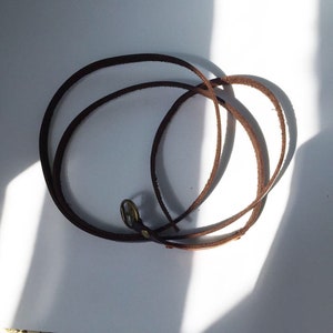 May include: A brown leather cord with a brass clasp. The cord is coiled and laid flat on a white surface.