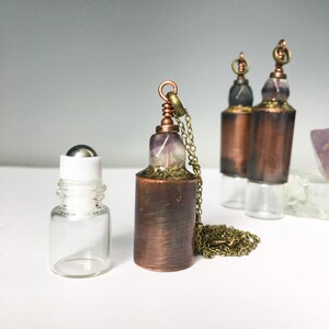 May include: Three copper and glass roller bottles with a silver roller ball. The bottles are filled with purple and white crystals. The bottles are on a white surface. Two of the bottles have a gold chain attached to the top.