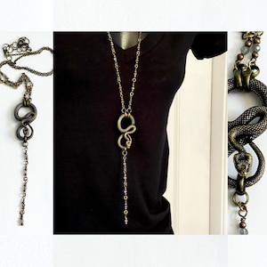 May include: A long, layered necklace with a snake pendant. The snake is made of metal and has a textured finish. The necklace is made of small, round beads and has a long, dangling chain.