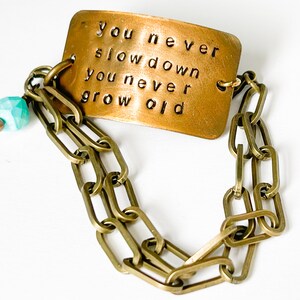 May include: A brass bracelet with a rectangular charm that reads "you never slow down you never grow old". The charm is attached to a chain with a turquoise bead.