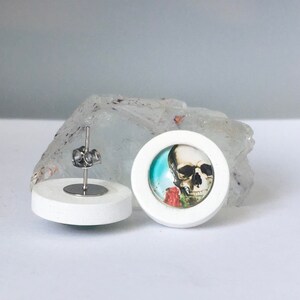 May include: A pair of white wood stud earrings with a skull and rose image in a glass cabochon. The earrings have stainless steel posts.