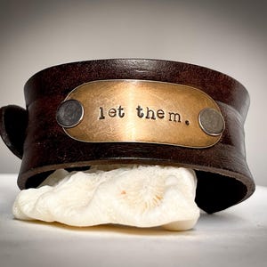 May include: A brown leather cuff bracelet with a brass plate that reads "let them." The plate is secured with two silver rivets.