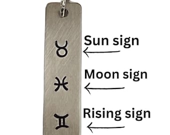 May include: A silver-colored rectangular pendant necklace with three astrological symbols: Taurus, Pisces, and Gemini. The text "Sun sign", "Moon sign", and "Rising sign" are next to the symbols. The pendant hangs from a delicate chain.