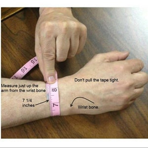 Op de afbeelding: Een roze meetlint is om de pols van een persoon gewikkeld om te laten zien hoe je voor een armband moet meten. De tekst op de afbeelding luidt "Meet gewoon omhoog langs de arm vanaf het polsbot 7 1/4 inch" en "Trek het lint niet te strak. Polsbot."