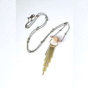 May include: A long, antique brass necklace with a geometric wooden bead and a cascade of brass bars. The necklace features a delicate chain with small, round beads and a lobster clasp closure. The pendant is suspended from a gold-toned ring.