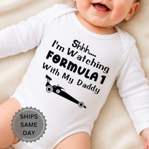 Könnte beinhalten: Ein weißer, langärmeliger Babybody mit schwarzem Text, der Shhhh.... I'm Watching FORMULA 1 With My Daddy lautet, begleitet von einer schwarzen grafischen Abbildung eines Rennwagens mit der Nummer eins an der Seite.