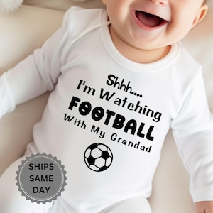 Op de afbeelding: Een wit babyrompertje met lange mouwen voorzien van de zwarte tekst Shhhh.... I'm Watching FOOTBALL With My Grandad en een afbeelding van een zwart-witte voetbal. Het kledingstuk heeft een ronde halslijn en lange mouwen voor volledige bedekking.