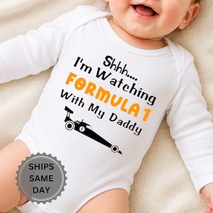 Könnte beinhalten: Ein weißer, langärmeliger Babybody mit schwarzem und orangefarbenem Text, der Shhhh.... I'm Watching FORMULA 1 With My Daddy lautet, begleitet von einer schwarzen grafischen Abbildung eines Rennwagens mit der Nummer eins an der Seite.