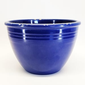 May include: A deep cobalt blue ceramic mixing bowl with a glossy finish and two horizontal ridges encircling the upper exterior. The thick, rounded rim shows visible signs of wear with small chips in the glaze along the top edge.
