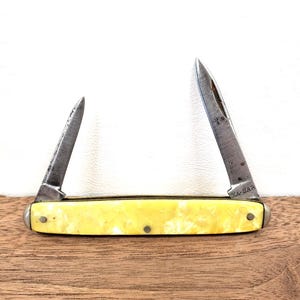 May include: A vintage pocket knife with two open blades. The knife features a yellow handle with a marbled pattern and metal rivets. The blades are silver and show signs of wear. The text "KA-BAR" is visible on one of the blades.
