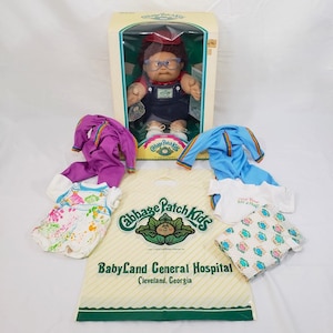 May include: A Cabbage Patch Kids doll in its original box, with several outfits and a shopping bag. The doll has brown hair, glasses, and is wearing overalls. The outfits include a purple tracksuit, a blue tracksuit, and two patterned shirts.