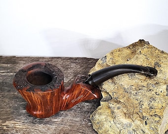 May include: A handcrafted tobacco pipe with a dark brown, curved stem and a reddish-brown bowl. The bowl has a textured, carved design around the rim. The pipe rests on a wooden surface.