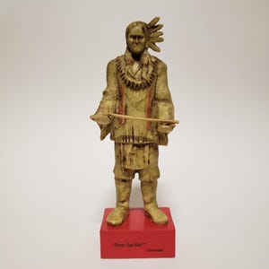 May include: A detailed figurine of a Native American figure in traditional attire, holding a spear. The statue is painted in shades of gold and brown, with red accents. It stands on a red base with the quote "Never Say Die!" and the name "Geronimo".