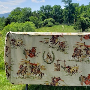 May include: A beige fabric featuring a repeating western-themed print with illustrations of cowboys on horseback, bucking bulls, lassos, and horseshoes. The scene depicts various rodeo activities and ranch life elements in shades of brown, red, and green.