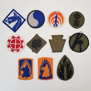 May include: A collection of embroidered military patches in various shapes and colors. Designs include a blue dragon, a yin-yang symbol, lightning bolts, dice, a four-leaf clover, and a stylized eagle with flames. The patches are likely for uniforms or collectibles.