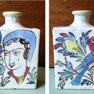 May include: A rectangular, white ceramic bottle with hand-painted designs. One side features a portrait of a person in a blue hat, the other a colorful bird with floral accents. The bottle has a small, rounded neck.