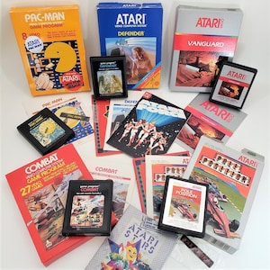 May include: A collection of Atari video game cartridges and boxes, including Pac-Man, Defender, and Combat. The boxes are in various colors, with game titles and graphics. Some cartridges are displayed with their instruction manuals.