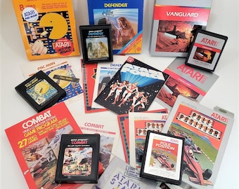 May include: A collection of Atari video game cartridges and boxes, including Pac-Man, Defender, and Combat. The boxes are in various colors, with game titles and graphics. Some cartridges are displayed with their instruction manuals.