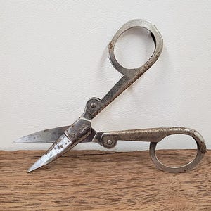 May include: Vintage metal scissors with finger loops. The scissors have a tarnished, aged appearance, with visible wear and tear on the blades and handles. The blades are pointed and sharp, and the scissor's hinge is visible.