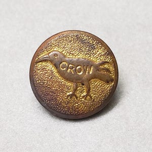 May include: A round, antique-style button with a crow design. The crow is in a side profile, with the word "CROW" above it. The button has a textured, gold-colored center and a darker, bronze-colored edge.