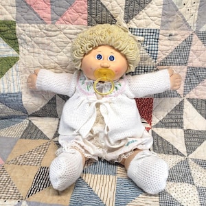 May include: A doll with blonde yarn hair and blue eyes, wearing a white knit cardigan with floral embroidery, a white ruffled shirt, and white knit socks. The doll has a yellow plastic pacifier in its mouth and is positioned with arms outstretched.