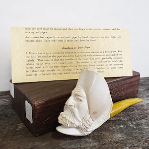 May include: A carved Meerschaum pipe with a detailed face design, featuring a yellow stem. The pipe rests on a wooden box, with a card providing instructions on breaking in the pipe. The text on the card reads "Breaking in Your Pipe."