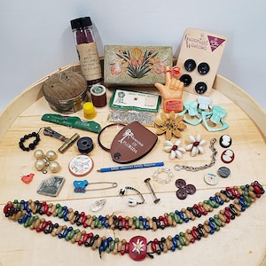 May include: A collection of vintage sewing notions and accessories, including a pincushion, thimble, spools of thread, buttons, and decorative pins. The items are arranged on a wooden tray, showcasing a variety of colors and textures. A few items have text, including "Souvenir of Florida".