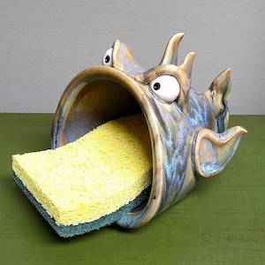 May include: A whimsical ceramic sponge holder shaped like a fish with wide eyes and an open mouth. The fish is glazed in shades of blue, brown, and gold. A yellow and green sponge is inside the fish's mouth. The holder is on a green surface.