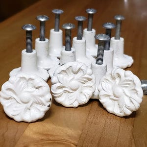 May include: A collection of white, floral-designed cabinet knobs with silver screws. The knobs have a distressed, vintage appearance, and are arranged on a wooden surface. The floral design is the focal point of the knobs.