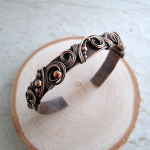 May include: A handcrafted copper cuff bracelet with intricate wirework and bead accents. The open-ended bracelet features swirling patterns and small copper beads, creating a unique, artistic design. The bracelet is displayed on a wooden surface.