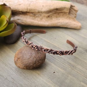 May include: A copper cuff bracelet with an open design. The bracelet features a decorative pattern of swirls and small beads. The jewelry is displayed on a brown stone, with a wooden background and a succulent plant in the background.