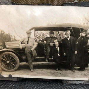 May include: A black and white vintage photograph of five people standing around a vintage car. The people are dressed in formal clothing and are smiling. The car is a touring car with a canvas top.