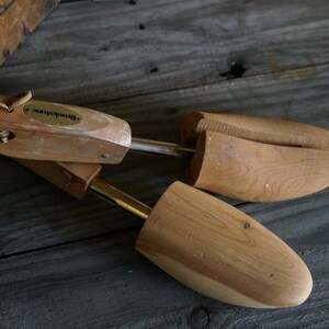 May include: A pair of wooden shoe trees with a brass metal rod connecting them. The shoe trees are made of light brown wood and have a label that says "Brooks Brothers".